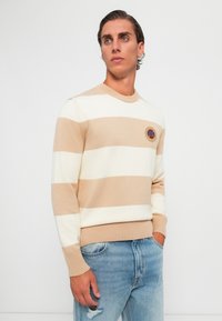 Randig tröja i beige och krämfärgad stickning, med rund halsringning och ribbade ärmslut. Inkluderar ett cirkulärt märke på bröstet.