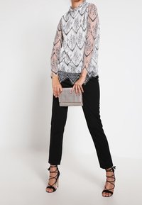 Top bianco trasparente con motivo nero, orlo scallop, abbinato a pantaloni neri e tacchi neri con cinturini. Tiene in mano una borsa a tracolla argentata.