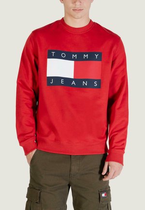Sweat-shirt rouge en tissu doux, avec un grand logo rectangulaire bleu marine et blanc portant le texte "TOMMY JEANS" au centre. Manches avec poignets côtelés.