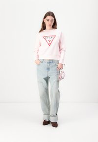 Sweat-shirt rose clair avec un logo triangulaire rouge et blanc, associé à un jean bleu clair ample et des bottes à talons marron, tenant un petit sac rose.