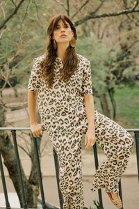 Kvinde med langt brunt hår, der bærer store runde øreringe og en leopardprintet outfit, der læner sig op ad et sort gelænder udendørs med træer i baggrunden.