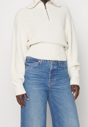 Persoon draagt een wit geribd vest met rits en oversized mouwen en een hoog uitgesneden, wijd uitlopende blauwe jeans, staand tegen een witte achtergrond.