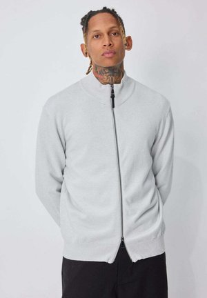Homme aux cheveux tressés et tatouage au cou, portant un pull à fermeture éclair gris clair et un pantalon noir, sur fond blanc uni.