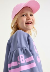 Paarse sweatshirts met opgerolde mouwen, voorzien van vet roze tekst en strepen. Een lichtroze baseballpet maakt de look compleet.