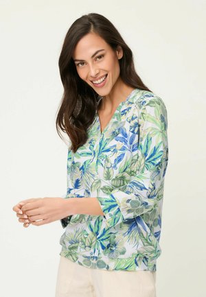 Donna sorridente con lunghi capelli scuri che indossa una camicetta bianca con motivi di foglie tropicali verdi e blu e pantaloni chiari.