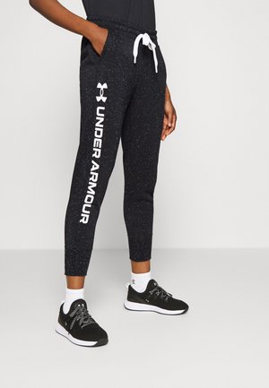 Person trägt schwarze, gesprenkelte Under Armour Jogginghosen mit weißem Kordelzug und schwarze Sportschuhe mit weißen Sohlen.