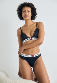 Zwarte kanten bralette met dunne bandjes en gekartelde randen; bijpassende thong met een witte merkband met rode en blauwe accenten.