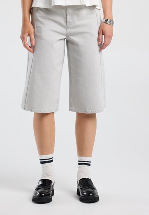 Persoon draagt lichtgrijze knielange shorts, witte sokken met zwarte strepen, zwarte loafers en een zilveren armband om de rechterpols.
