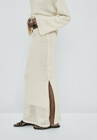 Personne portant une longue jupe en crochet crème avec une fente latérale, un pull assorti et des sandales plates noires, debout devant un fond uni.