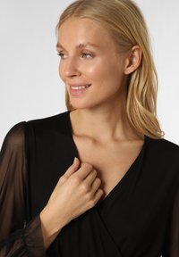 Schwarzes Kleid mit transparenten, gepufften Ärmeln, V-Ausschnitt und weicher Textur. Das Model lächelt und hält sanft den Ausschnitt.