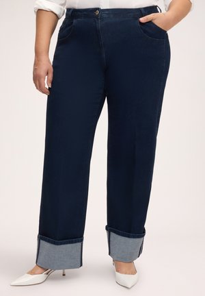 Person iført mørkeblå straight-leg jeans med kant og hvide spidse hæle med en hvid skjorteærme synlig.