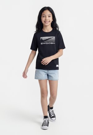 Chica caminando con camiseta negra de Nike Swoosh, pantalones cortos de mezclilla claros y zapatillas negras de caña alta sobre fondo blanco.