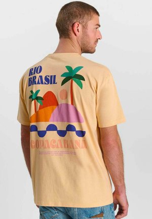 Man draagt beige T-shirt met kleurrijke afbeelding van palmbomen, heuvels en tekst "RIO BRASIL" en "COPACABANA" op de achterkant.