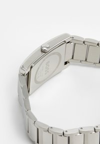 Relógio de pulso em aço inoxidável prateado com um estojo retangular, bracelete de elos metálicos e uma coroa. A parte de trás apresenta gravação da marca e detalhes sobre a resistência à água.