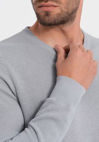 Grauer gestrickter V-Ausschnitt-Pullover mit gerippten Bündchen, der ein strukturiertes Muster und eine taillierte Passform aufweist, teilweise auf dem Oberkörper eines Models gezeigt.