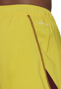 Gele actieve shorts met een elastische tailleband, contrasterende kastanjebrown zijpanelen en gestikte accenten met een logo aan de bovenkant.