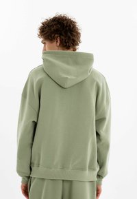 Grön överdimensionerad hoodie med huva, med ribbad mudd och fåll. Tillverkad av mjukt tyg med en slät yta. Visa bakifrån.