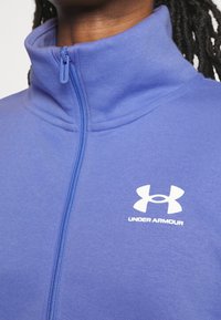 Chaqueta azul con cremallera y cuello alto. Hecha de un material suave, presenta un logotipo blanco de Under Armour en el lado izquierdo del pecho y una cremallera a juego.