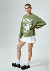 Olive zielony, oversize'owy sweter z białym nadrukiem graficznym, zestawiony z białą plisowaną spódnicą i czarnymi loafersami na platformie z złotymi elementami.