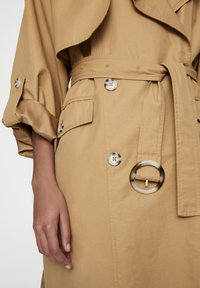 Tan trenchcoat met een ceintuur in de taille, grote knopen en een gelaagde kraag. Glad stof met een lichte glans; inclusief zijzakken.