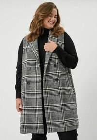 Dlhá károvaná vesta v čierno-bielej farbe, s klopovým golierom, dvojradovým zapínaním a voľným strihom, nosená cez čierny rolák.