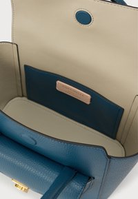 Blåt læderhåndtaskeinteriør med en glat, lys beige foring. Indeholder en lille lomme med et guld- og lyserødt mærke.