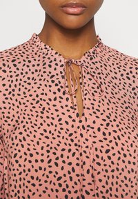 Světle růžová halenka s černým dalmatinským vzorem, s volánkovým výstřihem a zavazováním vpředu. Hladká textura látky.