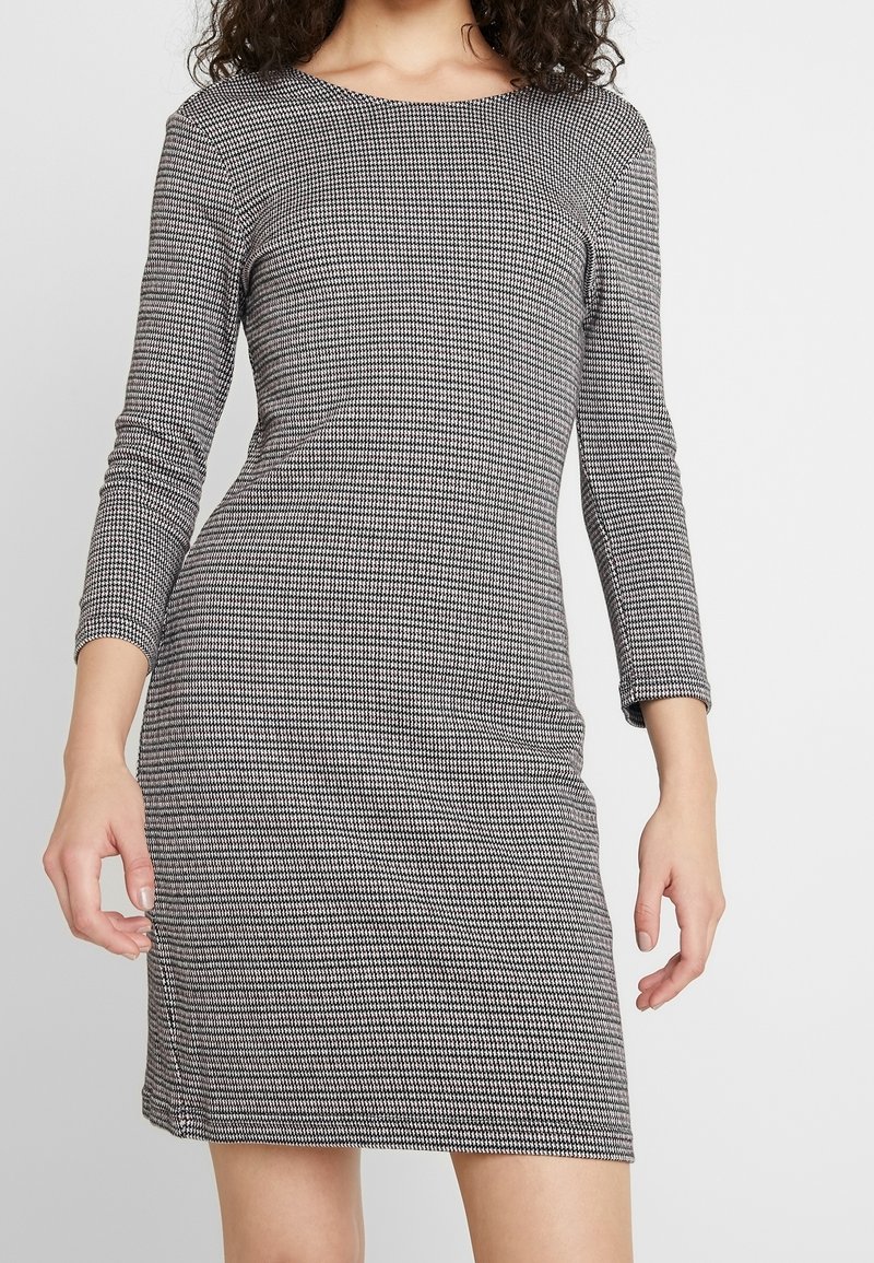 Femme portant une robe ajustée à la longueur genou avec des manches longues et un petit motif pied-de-poule noir et blanc, debout devant un fond uni.