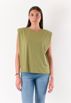 Donna con lunghi capelli scuri che indossa una canotta verde oliva con spalline imbottite e jeans azzurri chiari, in piedi contro uno sfondo bianco.