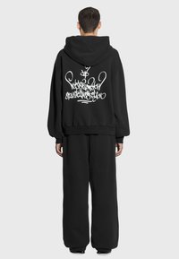 Czarna, oversize bluza z kapturem z dużym białym wzorem graffiti na plecach, zestawiona z dopasowanymi szerokimi czarnymi spodniami. Miękki, fakturowany materiał.