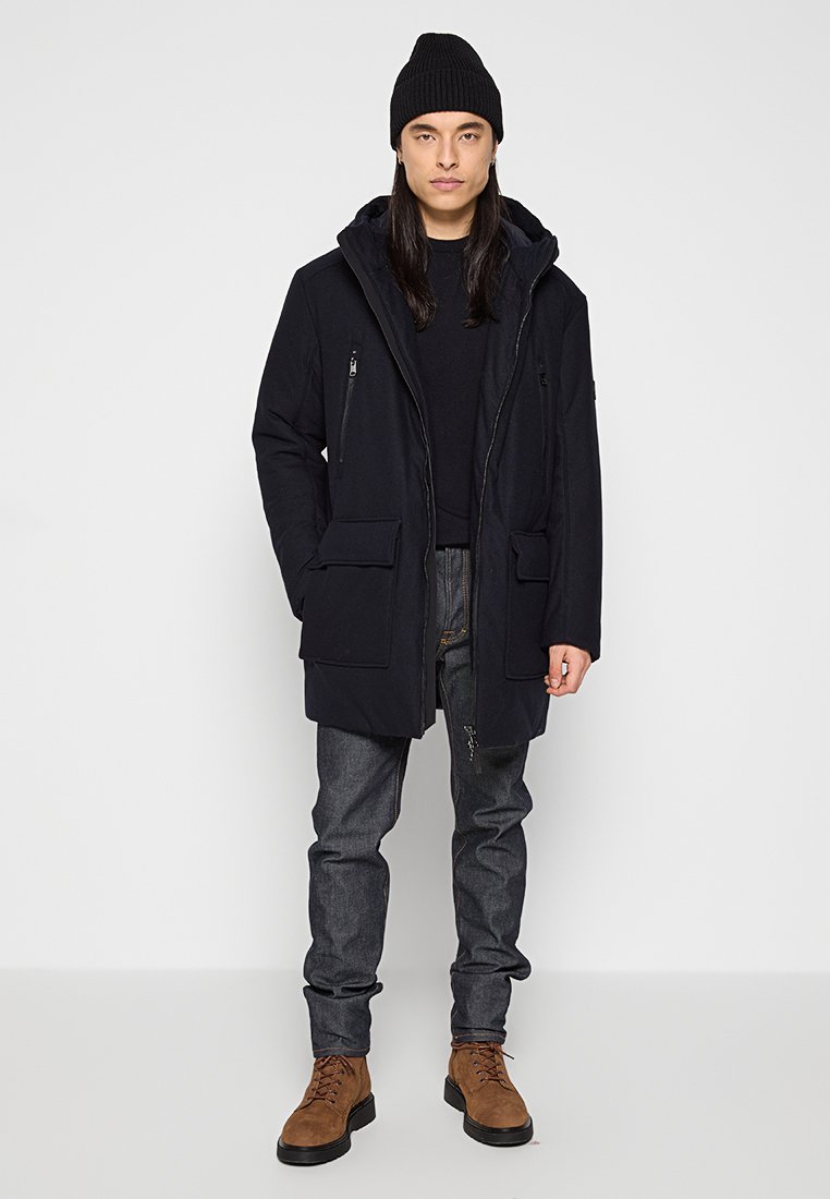 Schwarze Außenjacke mit Kapuze, mit einem Reißverschluss vorne, zwei großen vorderen Taschen und einem dezenten strukturierten Stoff. Kombiniert mit grauen Jeans und braunen Stiefeln.