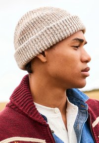 Beige ribbed knit beanie with light and dark fibers. Worn with a red and cream sweater and a denim shirt, showcasing layered textures.
