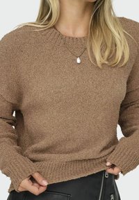 Maglione a maglia marrone con texture, colletto rotondo e maniche lunghe, abbinato a un pantalone in pelle nera con cerniere.