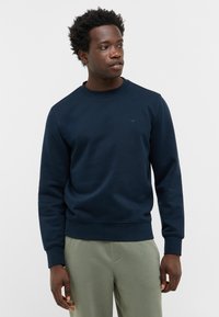 Sweatshirt bleu marine à col rond en tissu doux, présentant une finition texturée, des poignets côtelés et un petit logo sur la poitrine. Associé à un pantalon vert clair.