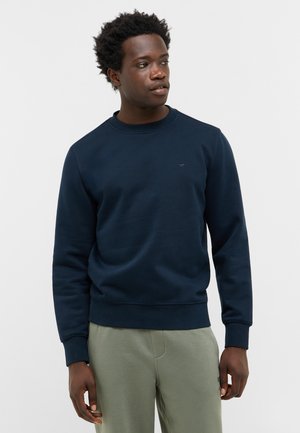 Marineblaue Rundhals-Sweatshirt aus weichem Material, mit strukturiertem Finish, gerippten Bündchen und einem kleinen Logo auf der Brust. Dazu leichte grüne Hose.