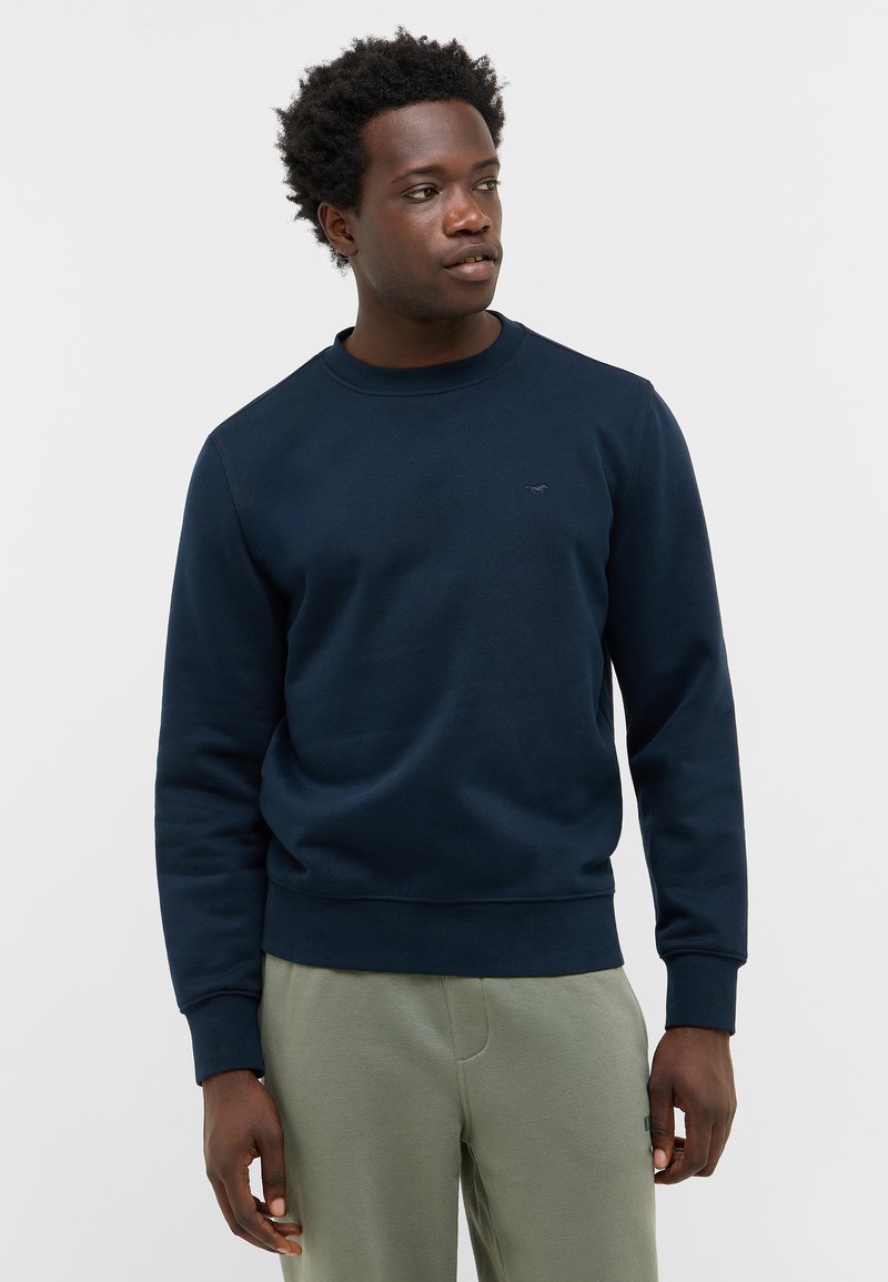 Sweatshirt bleu marine à col rond en tissu doux, présentant une finition texturée, des poignets côtelés et un petit logo sur la poitrine. Associé à un pantalon vert clair.