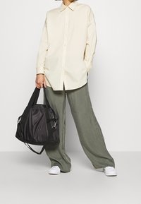 Sac à bandoulière en nylon noir avec doubles poignées et texture brillante, associé à une chemise boutonnée jaune clair et un pantalon ample vert.