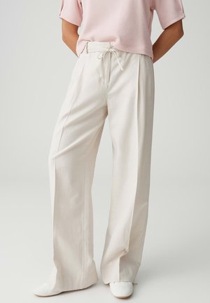 Femme portant un pantalon ample crème taille haute avec une ceinture à nouer, associé à des chaussures blanches et un haut à manches courtes rose pâle.