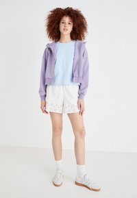 Lilla cropped hoodie, lyseblå top, hvide shorts med hulmønster, hvide sokker og lys grå sneakers med brune såler.