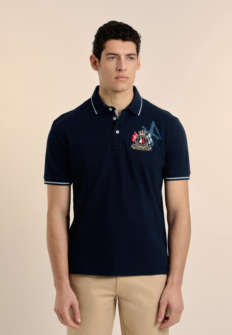 Polo navy con cuello, tres botones y acentos en azul claro en las mangas. Presenta un emblema decorativo en el pecho. Material de algodón.