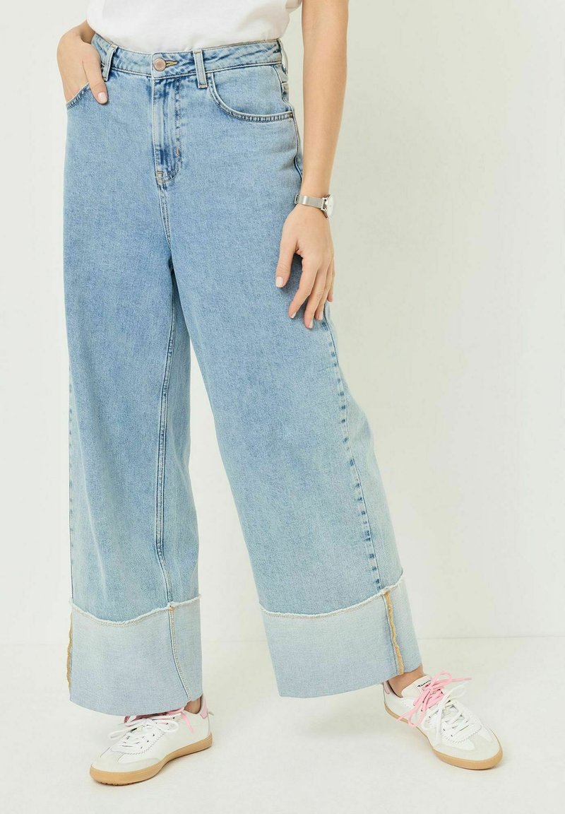 Personne portant un jean large bleu clair avec de grands revers et des baskets blanches aux lacets dépareillés rose et blanc.