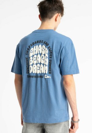 Młody mężczyzna w niebieskiej koszulce z napisem "CHASE BEACH DREAM" w białym i czarnym kolorze oraz dodatkowymi mniejszymi napisami na plecach.