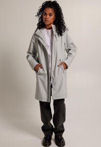 Light gray raincoat with a hood, front zipper, and side pockets. Made from waterproof material, features a relaxed fit and contrasting black pants.