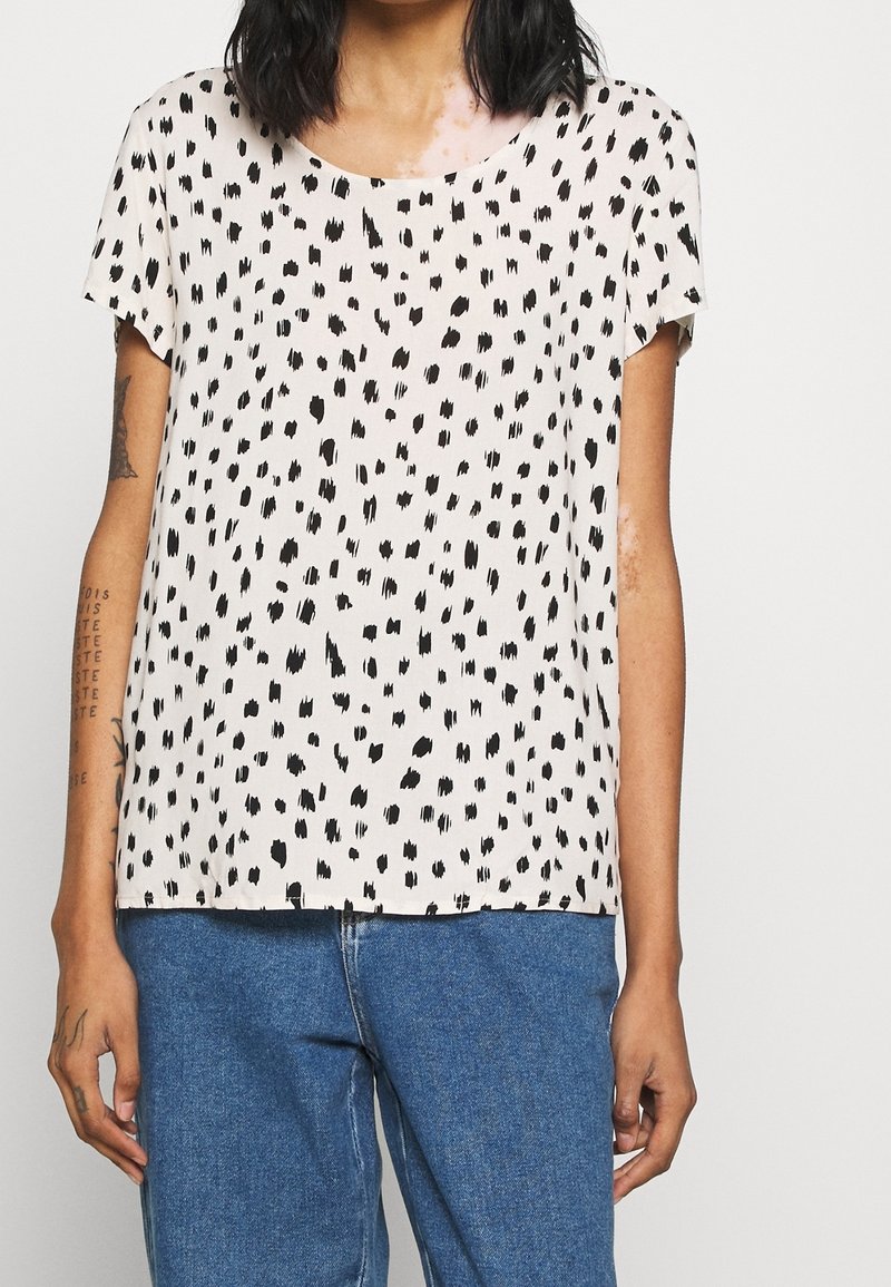 Personne portant une chemise blanche à manches courtes avec des taches noires irrégulières et un jean bleu, montrant des tatouages sur les bras.