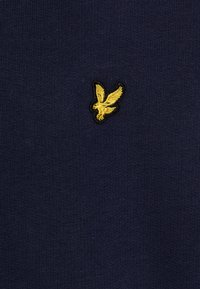 Marinblått stickat tyg med en liten, broderad gul fågel-logotyp, placerad på vänster sida strax nära bröstet.