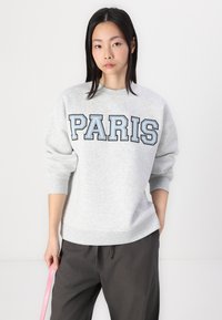 Felpa grigia chiara con la parola "PARIS" in lettere outline blu scuro. Caratterizzata da un collo tondo e polsini a coste, indossata con pantaloni scuri.