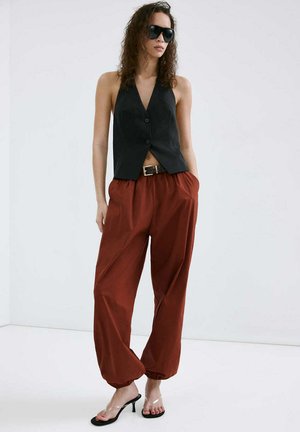 Femme portant un gilet noir sans manches boutonné, un pantalon ample couleur rouille, des sandales transparentes à talons et de grandes lunettes de soleil noires, debout les mains dans les poches.