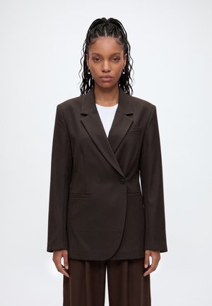 Femme aux cheveux tressés portant un blazer oversize marron foncé et un pantalon assorti, debout devant un fond clair uni.