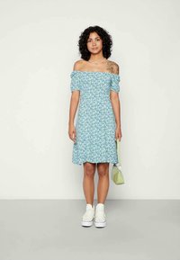 Mulher com cabelo encaracolado, vestindo um vestido azul floral off-shoulder, sapatilhas brancas, segurando uma pequena bolsa verde-claro, em pé contra um fundo simples.