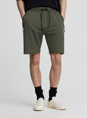 Uomo che indossa pantaloncini verde oliva con coulisse, calzini neri, scarpe da ginnastica bianche e maglietta nera con le mani in tasca.